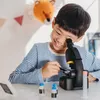 A young boy is happily using a microscope in a brightly lit room, with some decorative items in the background. Produktbild-Vorschau