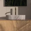 A beige matte washbasin with a gold faucet is shown in a bathroom setting, with a mirror reflecting greenery above. Produktbild-Vorschau