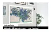 A framed television displaying a painting of blue irises, mounted on a wall with other framed pictures. A plant is visible to the right. Produktbild-Vorschau