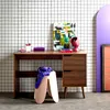 A modern desk setup with a wooden desk, a colorful abstract painting, a desk lamp, and a stool with a scarf draped over it, set against a grid-patterned wall. Produktbild-Vorschau