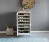 A white shoe cabinet filled with shoes, placed against a gray wall with a large clock above it. A woven basket is to the left of the cabinet. Produktbild-Vorschau