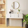 A round gold mirror hangs above a white console table with black legs, decorated with a lamp, clock, and plant. Produktbild-Vorschau