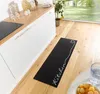A black kitchen runner with white text "Kitchen" and a heart design is placed on a wooden floor in a modern kitchen. Anteprima dell’immagine prodotto