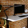 A black LED architect lamp is clamped to a white desk in front of a brick wall. A computer monitor and keyboard are also visible on the desk. Náhľad obrázku produktu