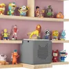 A collection of toys and a speaker are displayed on wooden shelves against a light purple wall. Produktbild-Vorschau
