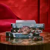 A holiday-themed advent calendar with a winter village scene is displayed on a reflective table, with a red velvet sofa in the background. Produktbild-Vorschau