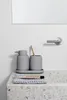 A gray ceramic soap dispenser and toothbrush holder sit on a gray tray on a speckled countertop next to a tube of toothpaste. Produktbild-Vorschau