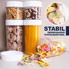 A collection of clear plastic food storage containers filled with various dry goods, including pasta, cereal, and colorful bow-tie pasta. Some containers are stacked, and one is tipped over, spilling its contents. Text overlays highlight product features. Produktbild-Vorschau