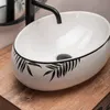 A close-up shot of a white oval sink with a black leaf pattern around the rim, placed on a wooden surface. Náhled obrázku produktu