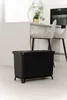 A black trash can with a lid sits on a light-colored floor in a modern kitchen setting. In the background, a white kitchen island with bar stools and a decorative plant is visible. Náhľad obrázku produktu