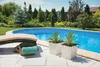 A backyard scene with a swimming pool, lounge chair, plants in pots, and a basket with a towel. Produktbild-Vorschau
