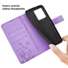 A hand is holding a black phone case over an open purple wallet-style phone case with card slots and a floral design. Produktbild-Vorschau