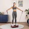 A woman is using a vibration plate machine with resistance bands in a living room. Náhled obrázku produktu