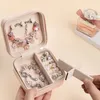 A person's hands are holding an open jewelry box filled with pink and silver bracelets and charms. A bottle of perfume is visible in the background. Produktbild-Vorschau