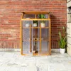 A wooden greenhouse with transparent panels, containing several potted plants, is placed on a tiled floor in front of a brick wall. Voir les images du produit