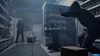Three people in a tense standoff inside a dimly lit store, possibly a convenience store or a warehouse. One person is on their knees with hands raised, another is standing with a weapon, and a third is in a tactical stance. Produktbild-Vorschau