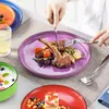 A person is eating a meal from a purple plate, with other colorful plates and food items visible. Produktbild-Vorschau