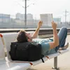 A man is lying on a bench at a train station, reading a book. Produktbild-Vorschau