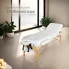 A white massage table is set up in a bright, modern room with large windows overlooking a body of water. Potted plants are placed around the room, and candles are visible in the foreground. Náhled obrázku produktu