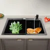A modern kitchen sink with a black basin, featuring a rack with green vegetables on the left and wooden bowls with soap dispenser and running water on the right. Tomatoes and a slice of papaya are visible on the countertop. Produktbild-Vorschau