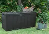 A dark gray Keter Sherwood garden storage box is placed on a green lawn in front of lush green foliage. A wooden crate labeled 'FLOWER' with gardening tools inside sits on top of the box. To the right, a potted plant in a metal bucket and a woven basket ar Anteprima dell’immagine prodotto