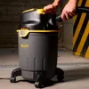 A person is holding the hose of a wet/dry vacuum cleaner. The vacuum cleaner is gray and black with yellow accents. The background is a concrete wall with yellow and black hazard stripes. Anteprima dell’immagine prodotto