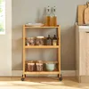 A three-tiered wooden serving cart filled with kitchen items like jars, plates, and bowls, placed against a gray wall. Voir les images du produit