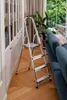 A silver step ladder is placed on a wooden floor next to a white cabinet and a blue sofa. A large green plant is in the background. Voir les images du produit