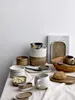 A collection of ceramic tableware, including mugs, bowls, and plates, arranged on a white tablecloth. Produktbild-Vorschau