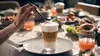 A person is stirring a layered coffee drink on a table set for breakfast. Anteprima dell’immagine prodotto
