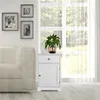 A white bedside table with one drawer and one door, placed next to a sofa and in front of a large window with sheer curtains. Produktbild-Vorschau