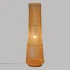 A tall, conical floor lamp made of thin, vertical rattan sticks, with a light bulb illuminated inside. Podgląd zdjęcia produktu