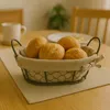A basket of bread rolls on a table, with a mug and a chair in the background. Anteprima dell’immagine prodotto