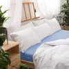 A neatly made bed with white pillows and a blue fitted sheet, with a wooden bedside table and plants. Náhled obrázku produktu