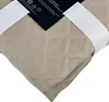 A close-up view of a taupe bedspread with a diamond pattern and small perforations, folded and held by white elastic bands. A product tag is visible in the upper left corner. Podgląd zdjęcia produktu