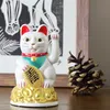 A white Maneki-neko cat figurine sits on a wooden surface, with a pine cone and a framed artwork in the background. Produktbild-Vorschau