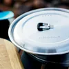 A close-up shot of a pot lid with a handle, resting on a stove. A wooden cutting board is visible to the left. Produktbild-Vorschau