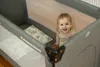 A baby is sitting in a gray travel crib with patterned pillows. Anteprima dell’immagine prodotto