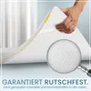 A hand is shown holding up a corner of a mattress topper, with a magnifying glass highlighting the textured underside. Produktbild-Vorschau