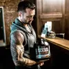 A man holding a large tub of protein powder in what appears to be a gym or fitness facility. Produktbild-Vorschau
