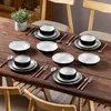 A set of tableware is arranged on a wooden table, with chairs visible in the background. Produktbild-Vorschau