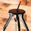 A close-up view of the top part of a black metal tripod stand with a hook and chain hanging from it. The background is a wooden wall. Produktbild-Vorschau