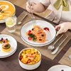 A person is eating pasta from a white plate with a black rim, with other dishes and cutlery on a wooden table. Náhled obrázku produktu