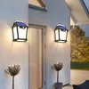 Two solar-powered wall lights illuminate a house exterior at dusk, with potted plants in the foreground. Produktbild-Vorschau