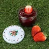 A jar of strawberry jam with a strawberry on top, a lid with a gingerbread man design, and two strawberries on the side, all placed on green grass. Produktbild-Vorschau