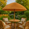 An outdoor patio set with chairs and a table under a thatched umbrella, surrounded by lush greenery and flowers. Voir les images du produit