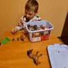 A young boy is playing with a set of animal figurines on a wooden table. A large bin of animal toys is next to him, and several figurines are scattered on the table. Anteprima dell’immagine prodotto