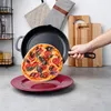 A hand is holding a cast iron skillet with a pizza inside, about to place it on a red plate. In the background, there are kitchen accessories. Produktbild-Vorschau