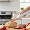 Weißes elektrisches Tranchiermesser, von einer Hand gehalten, schneidet ein Brot auf einem Holzschneidebrett mit Roastbeefscheiben, Tomaten, Knoblauch und Käse Anteprima dell’immagine prodotto