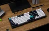 A keyboard is placed on a wooden desk, with glasses and other desk accessories visible. Produktbild-Vorschau